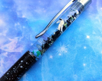 Op de afbeelding: Een zwart-zilveren pen met een ruimtethema. De doorzichtige behuizing bevat een miniatuur astronaut, een kleine aardbol en zwarte glitter. De pen heeft een zwarte behuizing met witte spikkels.