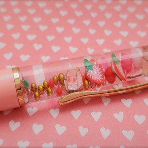 K&ouml;nnte beinhalten: Ein rosa und goldener Stift mit einem durchsichtigen Schaft, gef&uuml;llt mit bunten Perlen, Glitzer und kleinen Erdbeer-Anh&auml;ngern.
