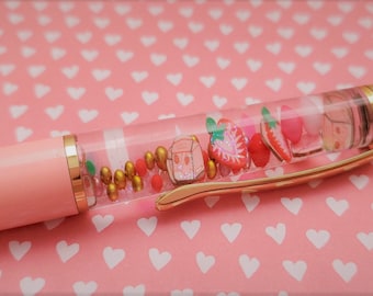 Op de afbeelding: Een roze en gouden pen met een transparante cilinder gevuld met gekleurde kralen, glitter en kleine aardbeiencharms.