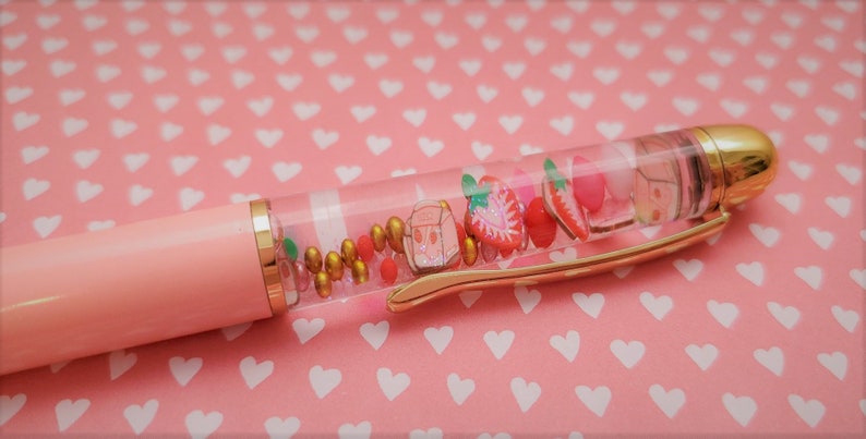 K&ouml;nnte beinhalten: Ein rosa und goldener Stift mit einem durchsichtigen Schaft, gef&uuml;llt mit bunten Perlen, Glitzer und kleinen Erdbeer-Anh&auml;ngern.