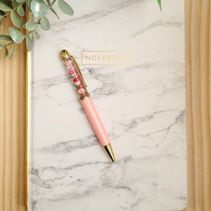 K&ouml;nnte beinhalten: Ein wei&szlig;es Marmor-Notizbuch mit goldfarbenem Rand und dem in Gold gedruckten Wort "NOTEBOOK". Ein rosa Stift mit einem durchsichtigen Schaft, gef&uuml;llt mit bunten Perlen, liegt auf dem Notizbuch.