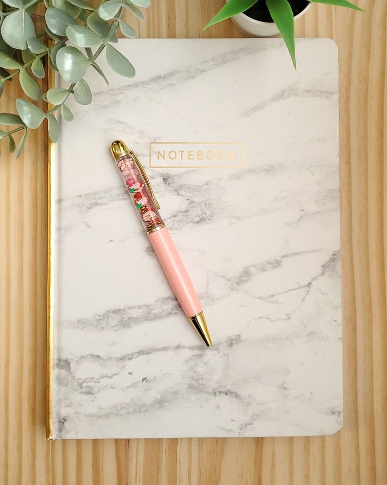 K&ouml;nnte beinhalten: Ein wei&szlig;es Marmor-Notizbuch mit goldfarbenem Rand und dem in Gold gedruckten Wort "NOTEBOOK". Ein rosa Stift mit einem durchsichtigen Schaft, gef&uuml;llt mit bunten Perlen, liegt auf dem Notizbuch.