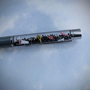 May include: A clear pen with a silver cap and a gray body. The pen contains small black beads, a yellow triangle, a pink ruler, and other small metal objects.