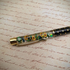 May include: A gold pen with a clear barrel filled with dried flowers and green glitter. The pen has a black cap with gold accents and small gold hearts.