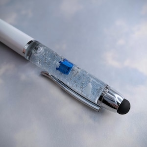 May include: A white pen with a clear body filled with silver glitter and a blue and white design. The pen has a silver tip and a black stylus.