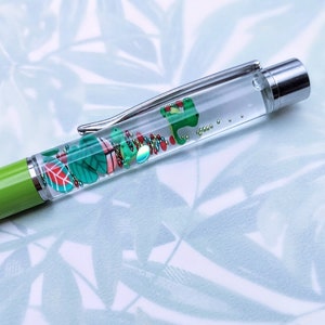 May include: A green and clear pen with a silver cap. The pen is filled with colorful glitter and small plastic shapes, including leaves and a frog.