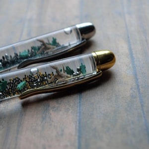May include: Two clear pens with gold and silver accents. The pens have a clear barrel filled with small green and gold beads and a small, brown, twig-like object.