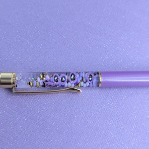 May include: A purple pen with a clear barrel filled with colorful glitter and small charms. The pen has a gold-colored clip and a gold-colored tip.