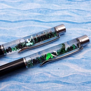 May include: Two black pens with clear barrels filled with green and black stones. The pens have silver accents.