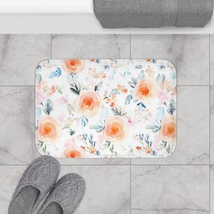 Könnte beinhalten: Eine weiße Badematte mit einem Blumenmuster mit pfirsichfarbenen Rosen und blauen Blättern. Die Matte liegt auf einem gefliesten Boden.