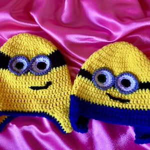 May include: Two yellow crochet hats with black stripes and blue trim, resembling Minion characters. Each hat features large, round eyes and a stitched smile. The hats are displayed on a pink satin surface.