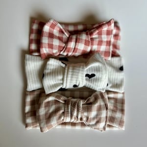 Puede incluir: Tres diademas acanaladas apiladas verticalmente. La diadema superior presenta un patrón de cuadros vichy en rosa empolvado y blanco. La diadema central es de color crema con pequeños corazones negros. La inferior tiene un patrón de cuadros vichy beige y blanco.