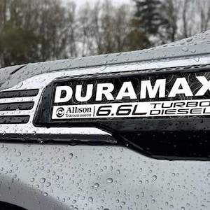 May include: A silver vehicle fender featuring a black emblem with the text DURAMAX, Allison Transmission, and 6.6L TURBO DIESEL. The emblem has a topographic line pattern background. The surface is covered in water droplets from rain.