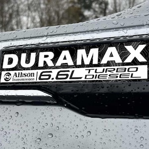 May include: A black and white Duramax Turbo Diesel emblem with the text "DURAMAX" and "6.6L TURBO DIESEL". The emblem also includes "Allison Transmission". The emblem is mounted on a silver vehicle panel with water droplets.