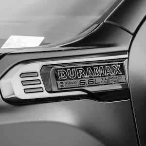 Può includere: Un emblema laterale per parafango di veicolo in nero con la scritta DURAMAX, 6.6L TURBO DIESEL e Allison Transmission. Il badge ha una finitura metallica con tre prese d'aria orizzontali sul lato sinistro, montato su un pannello della carrozzeria nero opaco.