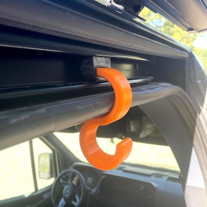 Op de afbeelding: Een oranje, C-vormige haak bevestigd aan een zwarte beugel, ontworpen voor gebruik in een voertuig. De haak is gemaakt van een fel oranje materiaal en hangt aan een zwarte stang. De binnenkant van het voertuig is zichtbaar op de achtergrond.