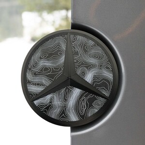 Könnte beinhalten: Ein schwarzes Emblem mit einem topografischen Kartenmuster in weißen Linien. Das Emblem hat die Form eines dreizackigen Sterns in einem Kreis, montiert auf einer grauen Oberfläche. Das Design ist detailliert und modern.