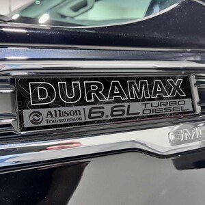 May include: Close-up of a black and chrome vehicle emblem. The emblem reads "DURAMAX" in white capital letters over a black topographic design. Below, it reads "Allison Transmission" and "6.6L TURBO DIESEL". The GMC logo is visible.