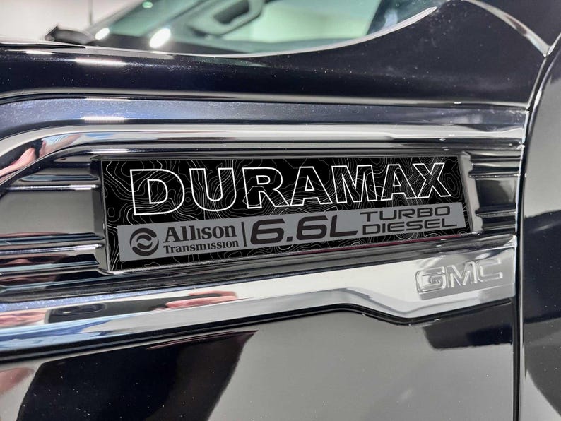 May include: Close-up of a black and chrome vehicle emblem. The emblem reads "DURAMAX" in white capital letters over a black topographic design. Below, it reads "Allison Transmission" and "6.6L TURBO DIESEL". The GMC logo is visible.