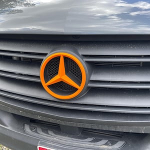 May include: Close-up of a dark gray vehicle grille with an orange and black emblem. The emblem features a three-pointed star within a circular frame. The grille has horizontal slats.