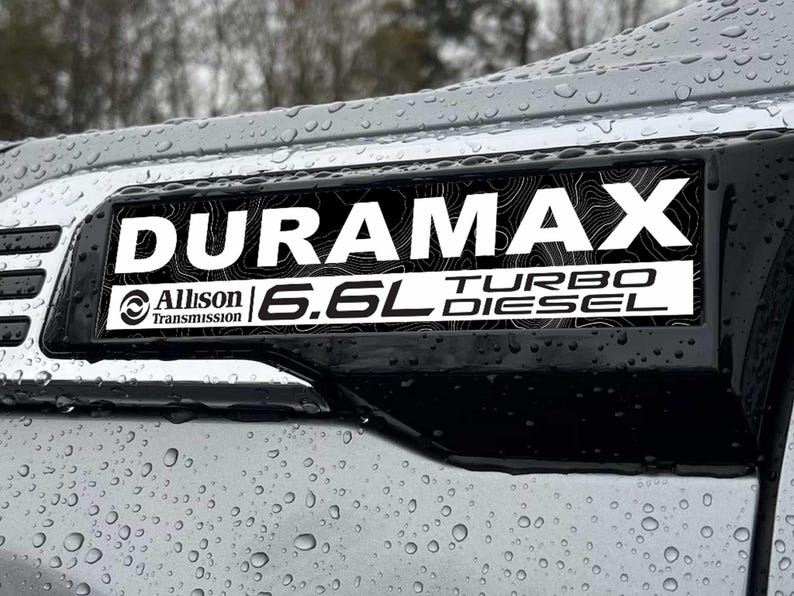 May include: A black and white Duramax Turbo Diesel emblem with the text "DURAMAX" and "6.6L TURBO DIESEL". The emblem also includes "Allison Transmission". The emblem is mounted on a silver vehicle panel with water droplets.