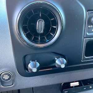 May include: Close-up of a black dashboard in a vehicle with a round air vent, two black knobs, and a button labeled "Start Engine Stop".