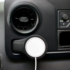 May include: A white wireless charging pad with a black rim is mounted to the dashboard of a car. The charging pad is connected to a white cable.