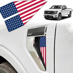 May include: Two rectangular decals featuring the American flag design with red and white stripes and a blue field with white stars. The decals are shown applied to the side vent of a white pickup truck, which also displays the text XLT on the trim.