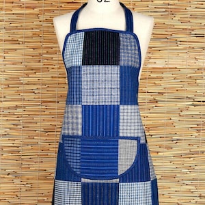 Puede incluir: Un delantal azul de patchwork con un bolsillo grande en la parte delantera. El delantal está hecho de diferentes telas azules y blancas con un patrón a cuadros.