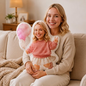 Könnte beinhalten: Ein individuell geformtes Dekokissen mit dem aufgedruckten Bild einer Person in einem rosa Pullover und einem hellen Rock, die ein Friedenszeichen zeigt. Das Kissen enthält eine Grafik mit rosa und weißen Luftballons auf der linken Seite.
