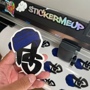 May include: A die-cut sticker featuring a stylized design with a blue turban and the letters "FS" in black. The sticker is held in a hand, with several similar stickers visible on a white surface. The text "STICKERMEUP" is visible.