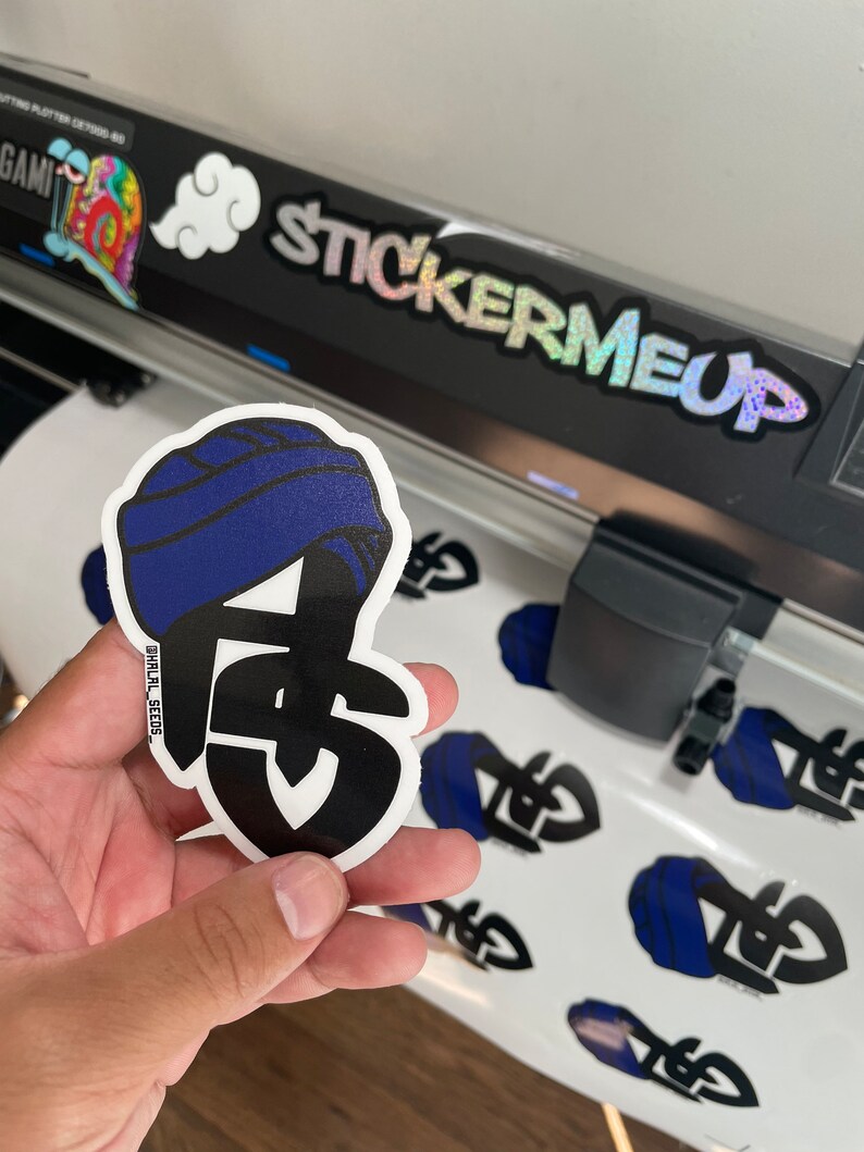May include: A die-cut sticker featuring a stylized design with a blue turban and the letters "FS" in black. The sticker is held in a hand, with several similar stickers visible on a white surface. The text "STICKERMEUP" is visible.