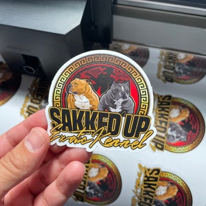 May include: A sticker with the text "SAKKED UP Exotic Kennel" in gold lettering. The sticker features two dog illustrations inside a red and black circle with a gold border. The sticker is held by a hand.