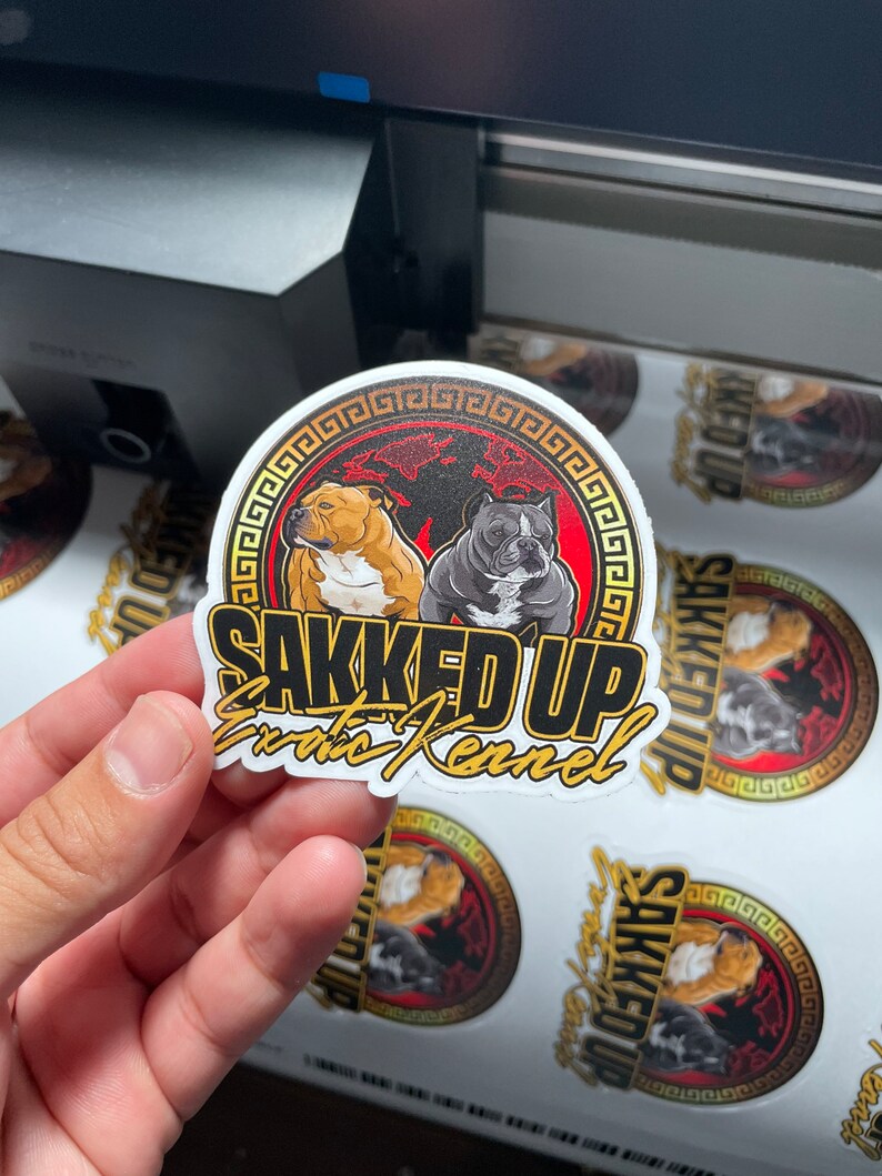 May include: A sticker with the text "SAKKED UP Exotic Kennel" in gold lettering. The sticker features two dog illustrations inside a red and black circle with a gold border. The sticker is held by a hand.