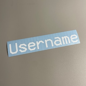 May include: A rectangular sticker with the word "Username" in white pixelated font on a light blue background. The sticker is on a gray surface.