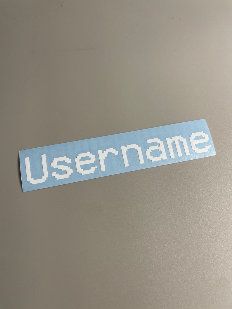 May include: A rectangular sticker with the word "Username" in white pixelated font on a light blue background. The sticker is on a gray surface.