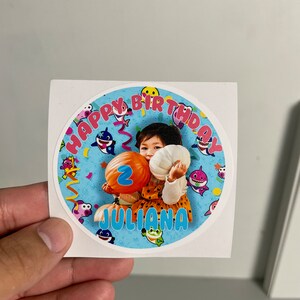 May include: A round sticker with a photo of a child holding a balloon with the number 2. The sticker has the text "HAPPY BIRTHDAY" and the name "JULIANA". The design includes cartoon sharks and confetti on a blue background.