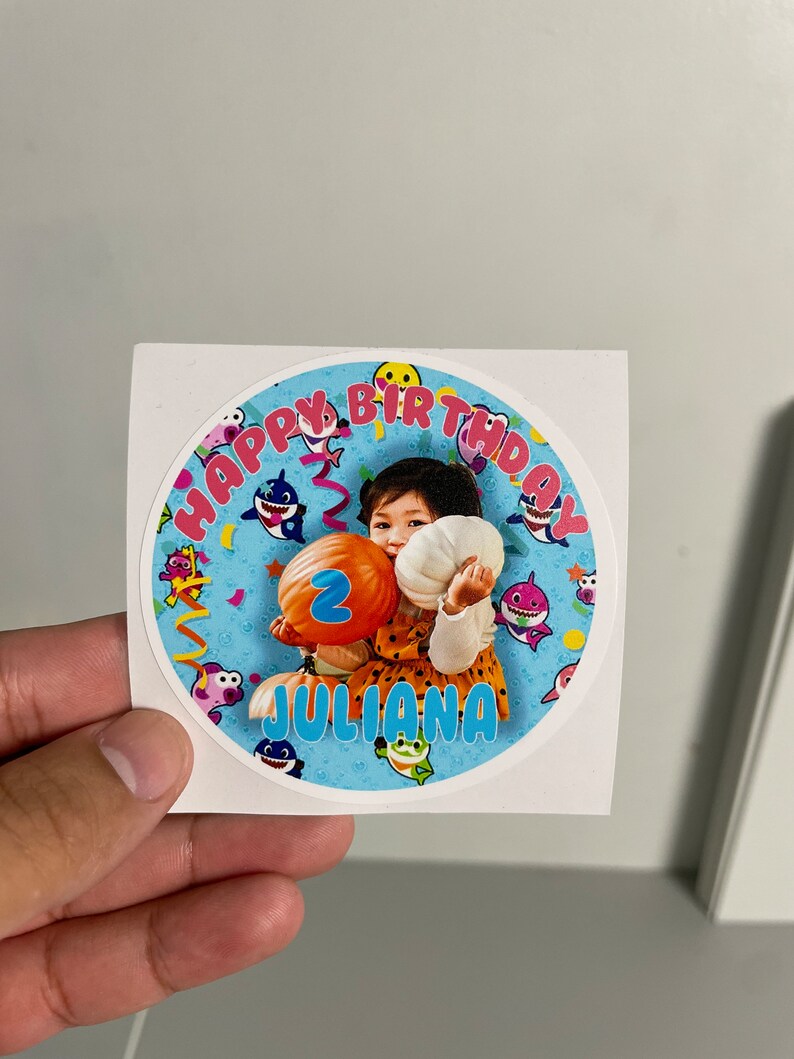 May include: A round sticker with a photo of a child holding a balloon with the number 2. The sticker has the text "HAPPY BIRTHDAY" and the name "JULIANA". The design includes cartoon sharks and confetti on a blue background.