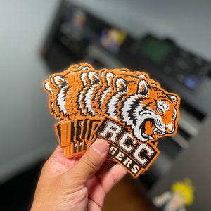 May include: A stack of orange and white tiger head stickers with the text "RCC TIGERS" in black and white. The stickers feature a detailed illustration of a tiger's face with orange and black stripes. The stickers are held in a hand.
