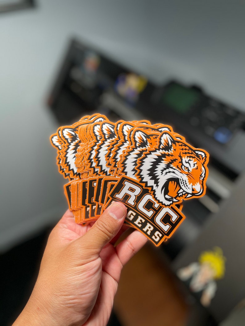 May include: A stack of orange and white tiger head stickers with the text "RCC TIGERS" in black and white. The stickers feature a detailed illustration of a tiger's face with orange and black stripes. The stickers are held in a hand.