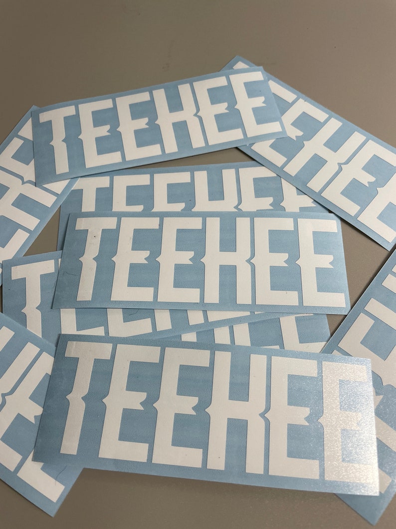 May include: A collection of rectangular stickers with the word "TEEHEE" in bold, white, serif font. The stickers have a light blue background. The stickers are scattered across a surface.