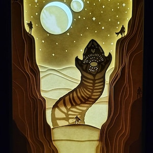 Peut inclure: Un diorama 3D en papier représentant un paysage désertique avec une grande créature vermiforme de couleur sable émergeant du sol. La créature a un corps texturé et segmenté et une grande gueule ouverte. Deux petites figurines se tiennent aux bords de la scène, regardant la créature. L'arrière-plan présente un ciel nocturne étoilé avec deux lunes.