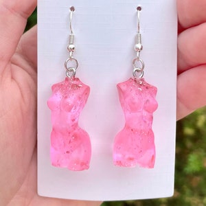 Puede incluir: Un par de pendientes de resina rosa con forma de torso femenino con purpurina incrustada en la resina. Los pendientes están en un gancho plateado.