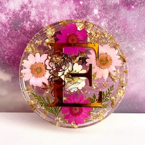 Op de afbeelding: Een ronde harsonderzetter met een gouden letter "E" omringd door geperste roze en witte bloemen en gouden bladaccenten.