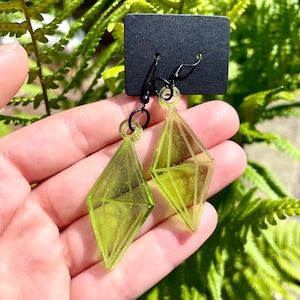 May include: A pair of lime green diamond-shaped earrings with black metal hooks. The earrings are hanging from a black card.