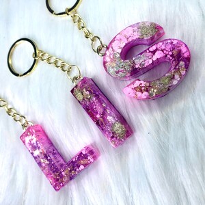 May include: Three gold keychains with pink and purple resin letters spelling "LIS" with gold glitter accents.