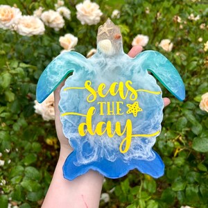 Pode incluir: Uma tartaruga de resina azul e branca com uma inscrição amarela que diz "Seas the day" e uma pequena estrela do mar e conchas incrustadas na resina.