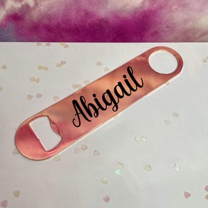 Puede incluir: Un abrebotellas de oro rosa con el nombre "Abigail" en escritura negra. El abridor tiene un cuerpo rectangular con un abridor de botellas en un extremo y un agujero circular en el otro.
