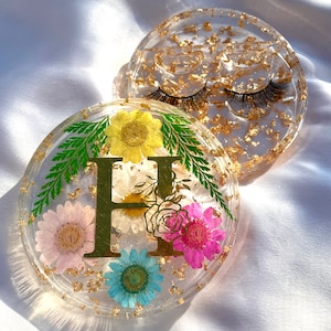 Op de afbeelding: Twee doorzichtige hars trays met goudfolie. Eén tray bevat valse wimpers. De andere tray heeft kleurrijke gedroogde bloemen, groene varen en een gouden letter "H". De bloemen zijn roze, geel en blauw.