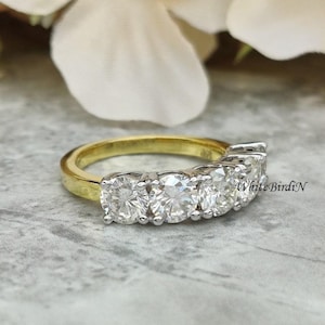 May include: A gold band ring with five round, clear gemstones set in a row. The ring is on a grey, textured surface.